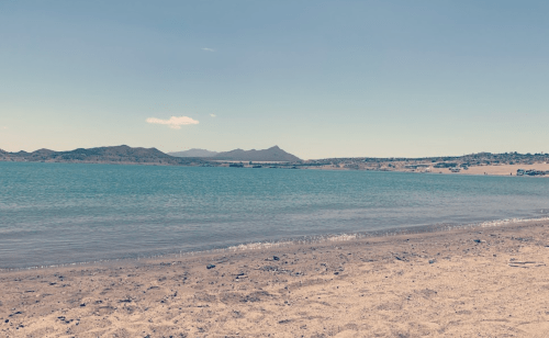 A serene beach scene with calm water, sandy shore, and distant mountains under a clear blue sky.