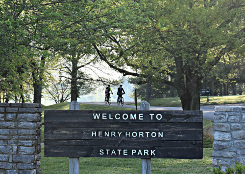Two cyclists ride past a wooden sign welcoming visitors to Henry Horton State Park, surrounded by lush greenery.