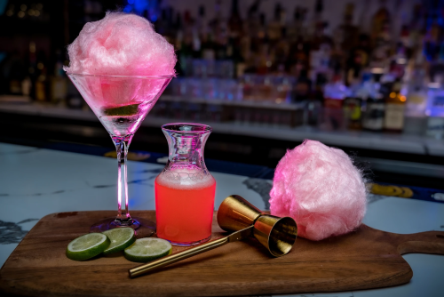 A colorful cocktail with cotton candy, a pink drink, lime slices, and a gold jigger on a wooden board.