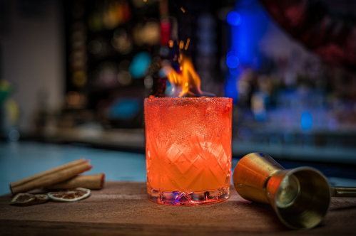 A vibrant cocktail with a flaming garnish, surrounded by cinnamon sticks and a jigger on a wooden bar.