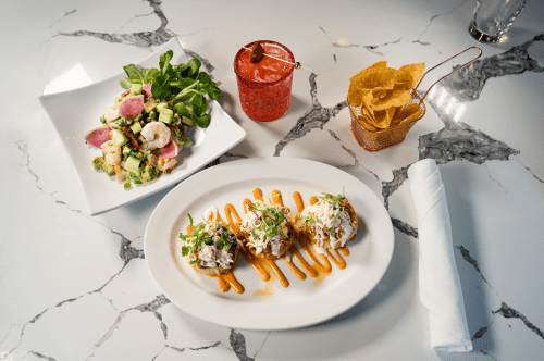 A plate with three tacos drizzled with sauce, a salad, a cocktail, and a basket of chips on a marble table.