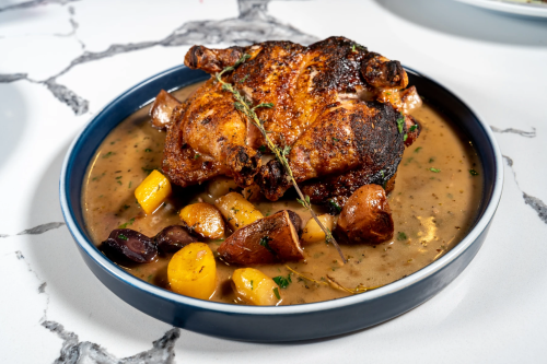 A roasted chicken on a plate with vegetables and gravy, garnished with herbs, on a marble surface.