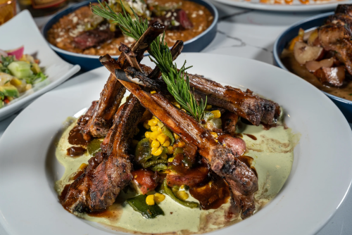 A plate of grilled lamb chops garnished with herbs, served on a bed of greens and corn, with sauces drizzled around.