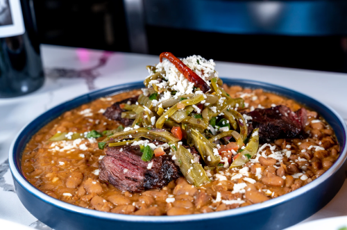 A plate of beans topped with grilled meat, sautéed peppers, and cheese, garnished with a red chili.