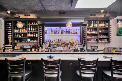 A stylish bar with a well-stocked shelf of bottles, modern lighting, and black chairs at the counter.