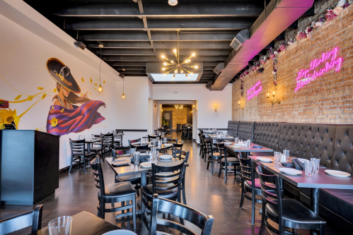 A modern restaurant interior featuring black chairs, a vibrant mural, and neon signs on exposed brick walls.