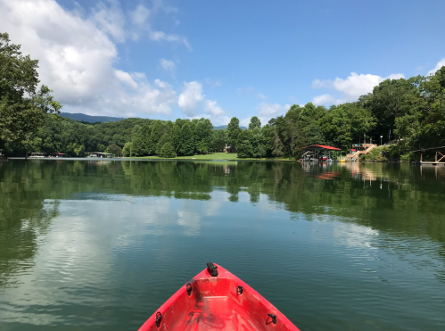 A red kayak on calm water, surrounded by lush green trees and a clear blue sky with fluffy clouds.