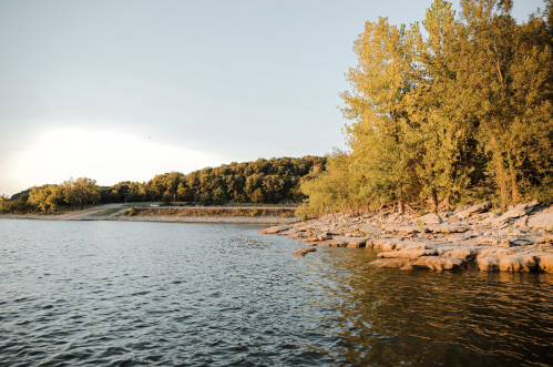 A serene lakeside view with rocky shorelines and trees reflecting golden sunlight under a clear sky.