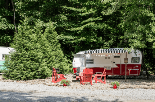 A vintage red and white camper parked among trees, with red chairs and potted plants in a gravel area.