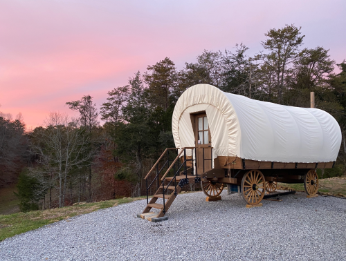 A covered wagon on a gravel area, surrounded by trees, with a colorful sunset sky in the background.