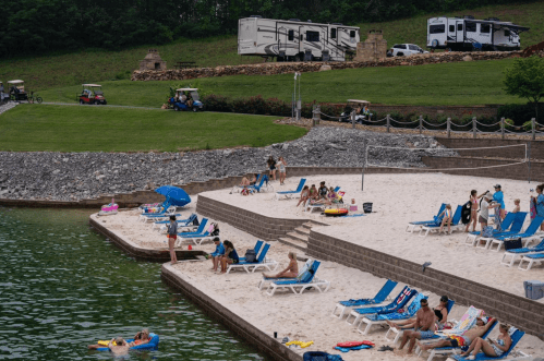 People relax on a sandy beach by a lake, with lounge chairs, umbrellas, and RVs in the background.