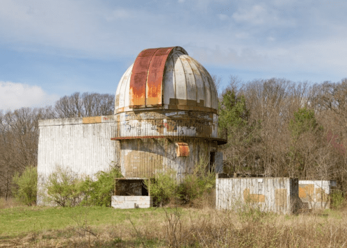 An abandoned observatory with a rusty dome, surrounded by overgrown grass and trees under a cloudy sky.