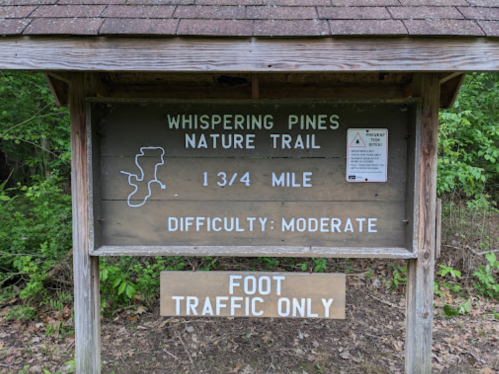 Sign for Whispering Pines Nature Trail, 1¾ mile, moderate difficulty, foot traffic only. Surrounded by greenery.