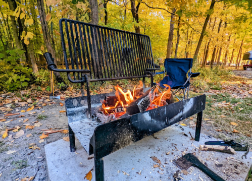 A campfire burns in a metal fire pit, with a folding chair nearby, surrounded by autumn trees with colorful leaves.