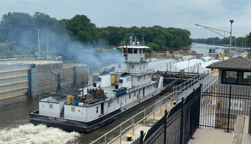 A towboat navigates a lock on a river, with smoke rising and a barge in tow, surrounded by green trees.