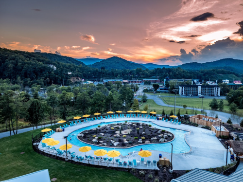 Aerial view of a circular pool surrounded by yellow umbrellas, with mountains and a sunset in the background.