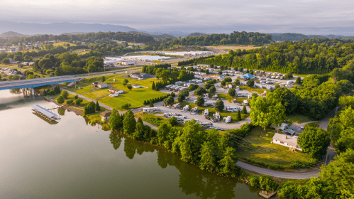 Aerial view of a lakeside RV park with green lawns, trees, and a bridge in the background.