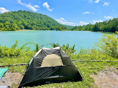 A tent by a serene lake surrounded by lush green hills under a clear blue sky.