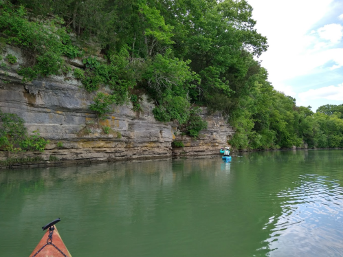 A serene river scene with a rocky cliff and lush greenery, featuring a person kayaking in the distance.