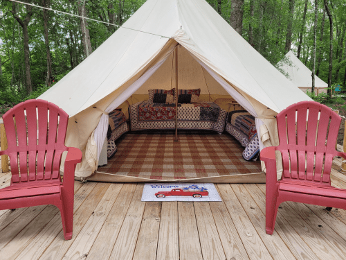 A cozy glamping tent with two red chairs outside, featuring patterned bedding and a welcome mat on a wooden deck.