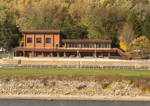 A modern building with large windows, situated by a river, surrounded by trees and a grassy area.