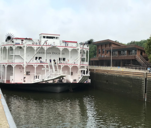 A large riverboat named "American Duchess" docked near a modern building by the water.