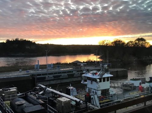 A river scene at sunset, featuring a boat docked near a lock, with colorful clouds reflecting in the water.