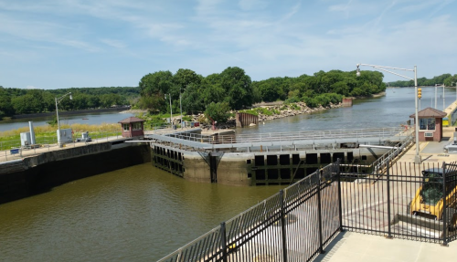 A view of a river lock with water flowing, surrounded by trees and a clear blue sky.