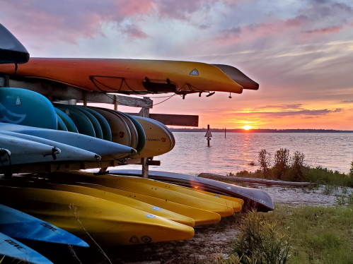Colorful kayaks stacked on a rack by the water, with a vibrant sunset in the background.