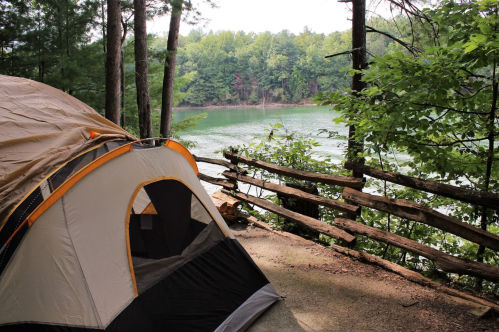 A tent by a serene lake, surrounded by trees and a wooden fence, creating a peaceful camping scene.