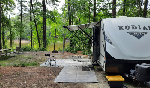 A camper parked by a serene lake, surrounded by trees, with outdoor chairs and a mat set up for relaxation.