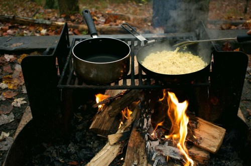 A campfire cooking setup with pots on a grill, one with boiling water and the other with steaming noodles.