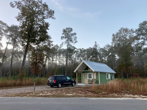 A small green cabin with a metal roof, parked black van, surrounded by trees and tall grass in a misty setting.