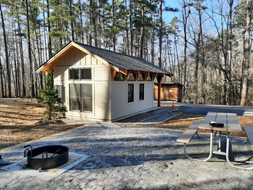 A small cabin surrounded by trees, with a picnic table and fire pit in the foreground on a gravel path.