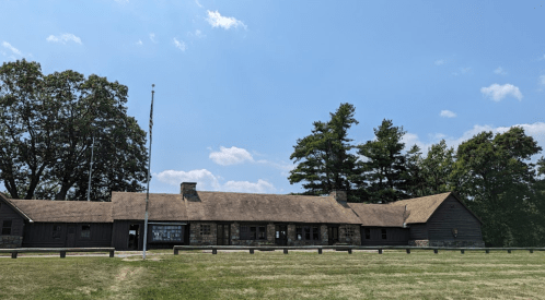 A large, rustic building with a thatched roof, surrounded by trees and a grassy field under a blue sky.