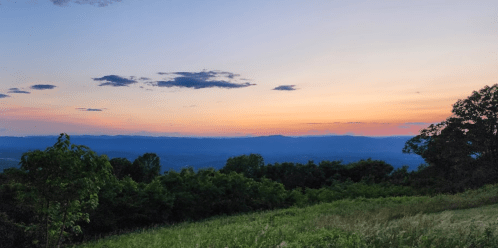 A serene landscape at sunset, featuring rolling hills, trees, and a colorful sky with soft clouds.