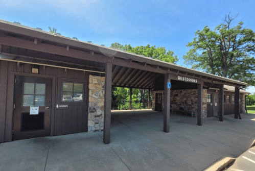 A rustic restroom building with stone accents and a covered entrance, surrounded by trees and blue sky.