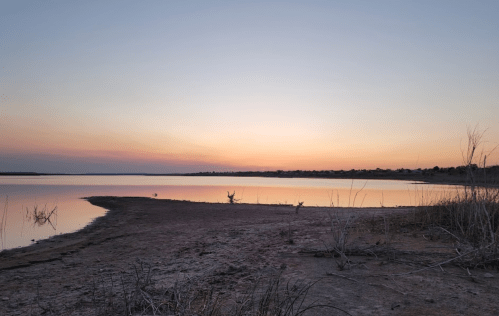 A serene lakeside view at sunset, with calm waters reflecting soft pastel colors and sparse vegetation along the shore.