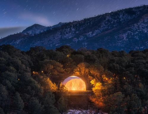 A glowing geodesic dome nestled in a forest, surrounded by mountains under a starry night sky.