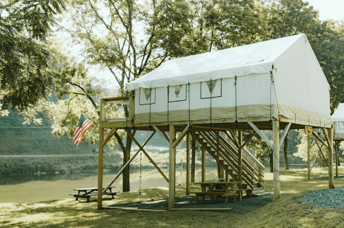 A raised tent cabin with a wooden deck, picnic table, and American flag, surrounded by trees and a calm river.