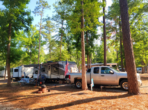 A camper and truck parked among tall trees in a wooded area, with outdoor seating and an American flag visible.