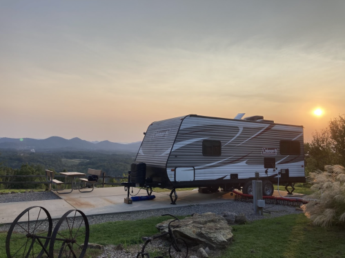 A camper parked on a scenic overlook with mountains in the background and a sunset casting a warm glow.