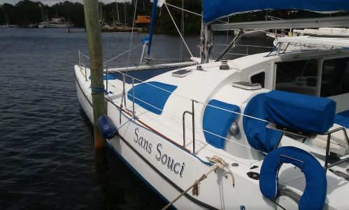 A white sailboat named "Sans Souci" docked at a marina, featuring blue accents and a calm water backdrop.