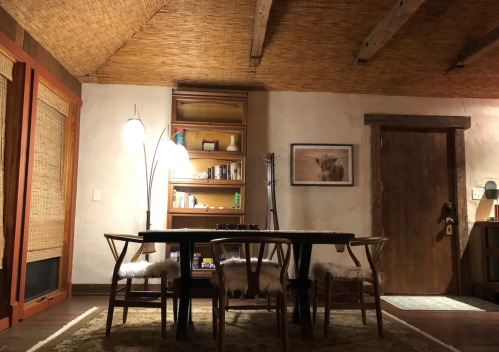 Cozy interior of a rustic room featuring a wooden table, chairs, a bookshelf, and warm lighting.