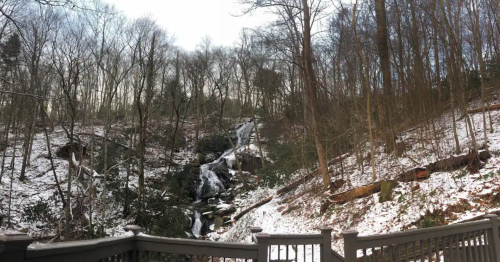 A snowy landscape featuring a waterfall cascading down a rocky slope, surrounded by bare trees and a wooden railing in the foreground.