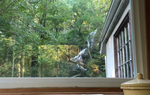 A view from a window showing a forested area with a waterfall and a nearby house.
