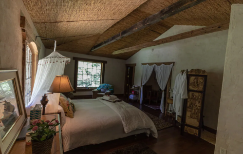 Cozy bedroom with a large bed, natural light, and rustic decor, featuring a lamp and a window with greenery outside.