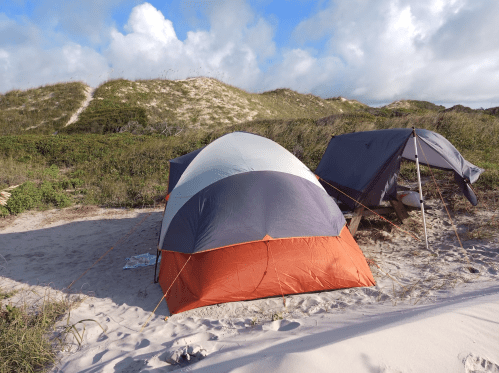 A camping scene featuring two tents set up on a sandy beach with dunes and cloudy skies in the background.