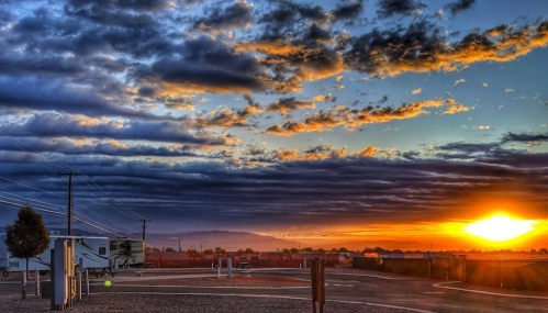A vibrant sunset over a landscape with clouds, power lines, and an RV park, casting warm colors in the sky.