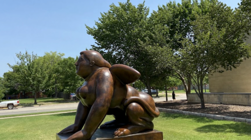 A large bronze sculpture of a seated, stylized figure with wings, surrounded by trees and a clear blue sky.